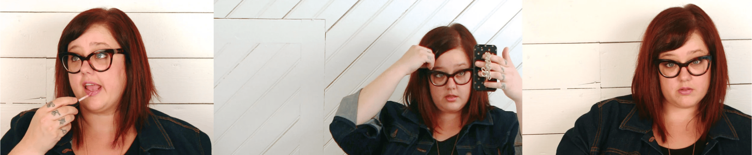 Woman with red hair and glasses in denim jacket applying lip gloss and taking mirror selfie against white paneled wall